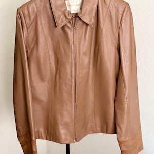 May include: A tan leather jacket featuring a pointed collar, a full-length front zipper closure, and long sleeves with horizontal panel detailing at the cuffs. The interior label displays the text Barneys New York in a white and gold font.