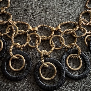 May include: A statement necklace featuring a series of interlocking gold-toned textured metal rings and larger black circular rings. The design consists of multiple connected loops arranged in a layered pattern, creating a bold, geometric aesthetic.