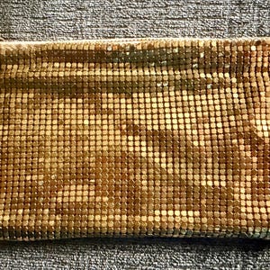 May include: A rectangular gold-colored metal mesh pouch featuring a fine, interlocking square-link pattern. The clutch has a smooth, reflective surface texture and includes a small, circular metal ring attached to the top corner for a zipper pull.