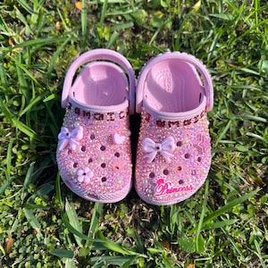 Pode incluir: Um par de Crocs rosa claro decorados com strass brilhantes. Os sapatos apresentam a palavra "Jamaica" em vermelho e um gráfico "Princess". Adornados com laços e flores, estão sobre a relva verde.