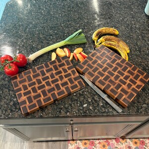May include: Two rectangular wooden chopping boards featuring a dark and light brown basketweave pattern. The boards are arranged on a dark countertop alongside three red tomatoes on a vine, a green leek, sliced apples, and three spotted yellow bananas.