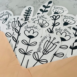 May include: A collection of white floral stickers with black outlines and text. The stickers include the phrases "Rosaries: 9", "Novenas: 1", "Holy Hours: 1/2", "Our Fathers: 5", "Chaplets: 1", "Memorares: 3", and "Fasting: 1".
