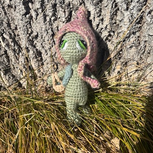 Könnte beinhalten: Handgemachte Häkelpuppe mit grünem Körper, großen grünen Augen und einer rosa-beige Kapuze. Die Puppe hat einen kleinen blauen Schal und steht im Gras. Der Hintergrund ist ein Baumstamm.