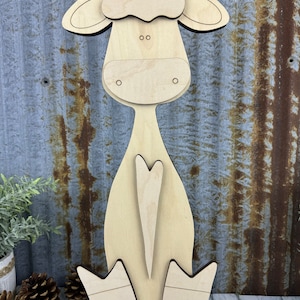 May include: A wooden cutout of a cow in a sitting position. The cow is light brown and has a white patch on its chest. The cow has a heart-shaped tail and large ears.