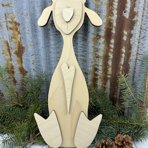 May include: A wooden cutout of a sheep with a heart on its chest. The sheep is standing on a white surface with pine branches and pine cones.