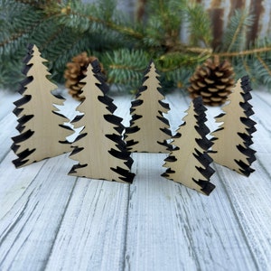 Peut inclure: Cinq décorations de sapin de Noël en bois avec un contour noir. Les décorations sont disposées en rangée sur une surface en bois blanc.