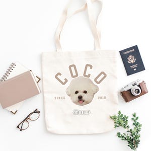 May include: White canvas tote bag with brown text that reads "Coco Since 2018" and a white dog's head in the center. The dog has a pink nose and tongue.
