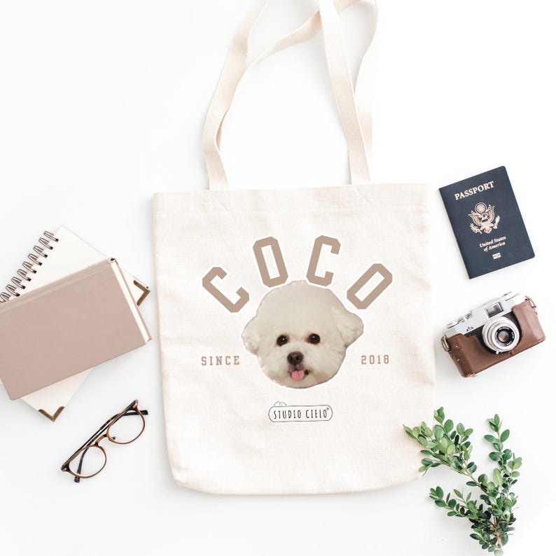 May include: White canvas tote bag with brown text that reads "Coco Since 2018" and a white dog's head in the center. The dog has a pink nose and tongue.