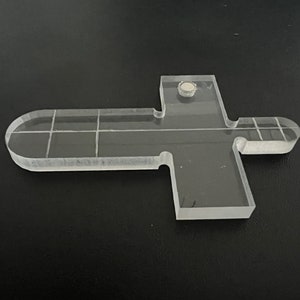Pu&ograve; includere: Un righello trasparente a forma di croce in plastica con un motivo a griglia e un piccolo magnete rotondo nella parte superiore.
