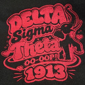 May include: A black fabric surface featuring red embroidery with the text Delta Sigma Theta, OO-OOP!, and 1913. The design includes a stylized elephant figure, small heart shapes, and star accents, all rendered in a bold, thick red thread outline.