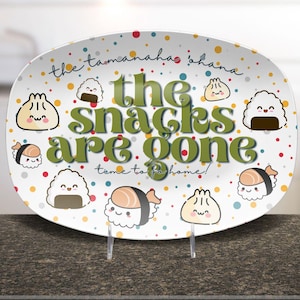 May include: White oval serving platter with the text "the snacks are gone" in green, surrounded by colorful polka dots and cartoon sushi and dumpling illustrations. The platter is propped up on a clear stand.