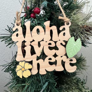 May include: Wooden ornament with the phrase "aloha lives here" in a retro font. The ornament features a yellow flower and a green leaf accent. It is hanging on a Christmas tree.