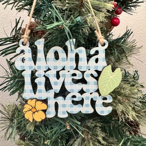 May include: Wooden ornament with the phrase "Aloha lives here" in blue and white checkered letters. A yellow flower and a green leaf accent the design. The ornament hangs from a natural twine string, displayed on a Christmas tree.