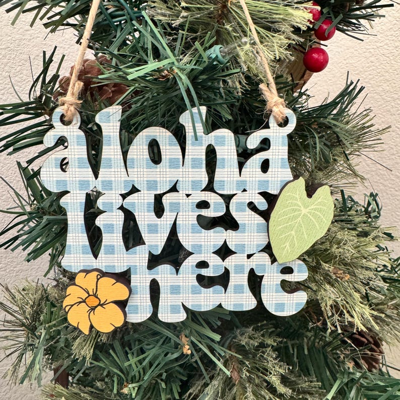 May include: Wooden ornament with the phrase "Aloha lives here" in blue and white checkered letters. A yellow flower and a green leaf accent the design. The ornament hangs from a natural twine string, displayed on a Christmas tree.