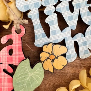 May include: Wooden cutouts spelling "SPRING" in blue and pink plaid patterns. The image includes a yellow flower, a green leaf, and yellow floral accents, all on a brown wooden surface.