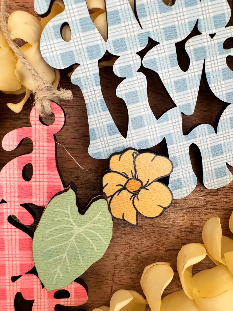 May include: Wooden cutouts spelling "SPRING" in blue and pink plaid patterns. The image includes a yellow flower, a green leaf, and yellow floral accents, all on a brown wooden surface.