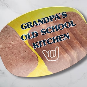 May include: An oval-shaped serving platter with a yellow background featuring a photographic print of sliced luncheon meat. The text GRANDPA'S OLD SCHOOL KITCHEN is printed in dark blue, accompanied by a white line-art shaka hand gesture icon.