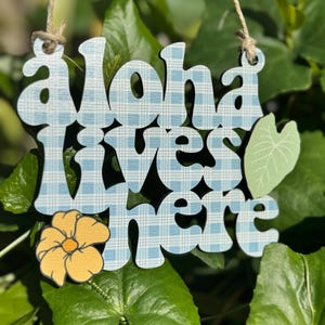 May include: Wooden sign with the words "Aloha lives here" in a blue plaid pattern. A yellow flower and a green leaf accent the sign, which hangs from a natural twine string. The sign is set against a backdrop of green leaves.