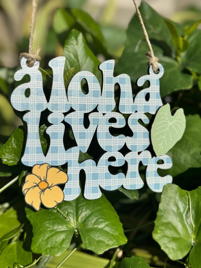 May include: Wooden sign with the words "Aloha lives here" in a blue plaid pattern. A yellow flower and a green leaf accent the sign, which hangs from a natural twine string. The sign is set against a backdrop of green leaves.