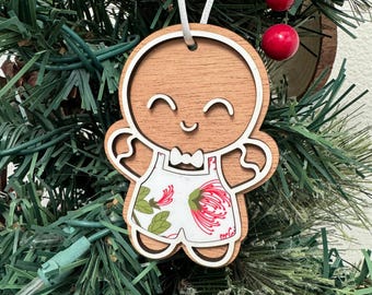 May include: Wooden gingerbread person ornament with a white apron and red flowers. The ornament is smiling and has a bow tie.