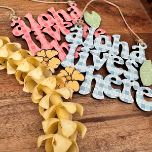 May include: Wooden sign with the words "Aloha Lives Here" in pink and blue plaid patterns, with yellow flower and green leaf accents. A lei made of yellow petals hangs below the sign. The sign is suspended by a natural twine.