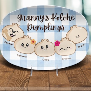 May include: An oval ceramic platter with a blue and white checkered pattern. The platter features the text "Granny's Kolohe Dumplings" and illustrations of five dumpling characters, each with a name.