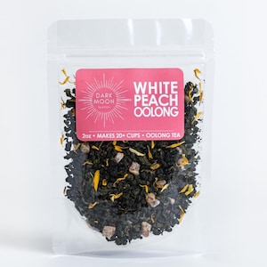 May include: A clear plastic pouch containing a blend of loose leaf tea. The tea is a mix of green tea, peach pieces, and yellow flower petals. The pouch has a pink label that reads "White Peach Oolong" and "Dark Moon Teaco." The label also states "2oz - Makes 20+ Cups - Oolong Tea."