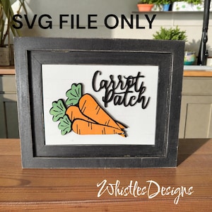 May include: A framed wooden sign with a white background, featuring the words "Carrot Patch" in black script above two orange carrots with green tops. The frame is dark gray, and the sign is on a wooden surface.