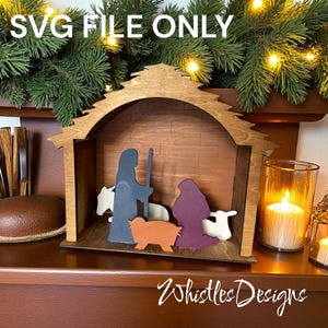 Puede incluir: Un belén de madera con un establo marrón y recortes coloridos de María, José, un cordero y otros animales. El texto "SVG FILE ONLY" está en la parte superior y "Whistles Designs" en la inferior. La escena está sobre una superficie de madera.