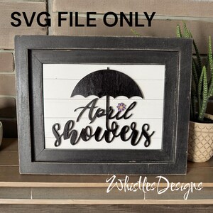 May include: A framed wooden sign with the text "April showers" in a cursive font, under a black umbrella. A small purple flower is next to the word "showers". The sign is on a white background with horizontal wooden slats.