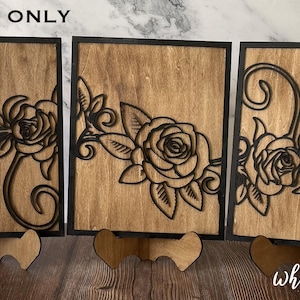 Może przedstawiać: Trzy drewniane dekoracje ścienne w czarnych ramach. Każdy element zawiera laserowo wycięty wzór róży z liśćmi i winoroślą. Elementy są prezentowane na małych drewnianych podstawkach.