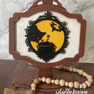 May include: A brown wooden frame with a black silhouette of a cat and a cauldron under a yellow moon. The frame is on a tiered stand with a wooden bead garland and a brown tassel.