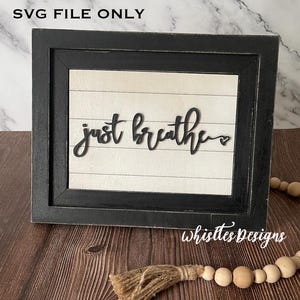 May include: Black framed sign with white background and the text "just breathe" in black script font with a small heart symbol. The sign is on a wood surface.
