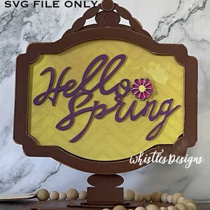 Puede incluir: Un letrero de madera con un marco marrón y un fondo amarillo con un patrón de diamantes. El letrero dice "Hello Spring" en letras moradas con una flor rosa.