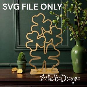 May include: A gold-colored, laser-cut shamrock decoration with the text "SVG FILE ONLY" at the top. The shamrock is on a wooden base. A small potted plant and a green vase with greenery are also in the image. The text "Whistles Designs" is at the bottom.