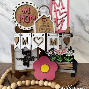 May include: A wooden tiered tray with a pink flower, a sign that says "Happy Mother's Day", and a banner that says "MOM". The tray is decorated with wooden beads and a house-shaped sign that says "Thanks for being our home".