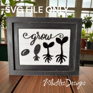 May include: A framed wooden sign with the word "grow" in cursive script. Below the word are three black cutouts depicting the stages of plant growth, from seed to sprout. The frame is black, and the background is white with horizontal lines.