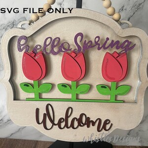 May include: A wooden sign with a white background and a decorative frame. The sign says "Hello Spring" in purple glitter letters and "Welcome" in brown letters. There are three red tulips with green leaves below the text.
