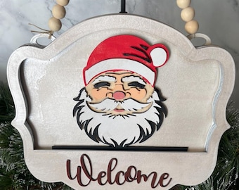 Puede incluir: Un letrero de madera blanco con una cara de Papá Noel roja y blanca y la palabra "Bienvenido" en letras rojas. El letrero está colgado de una guirnalda de cuentas.