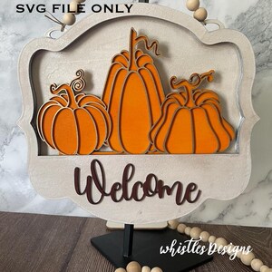 Könnte beinhalten: Holzschild mit drei detaillierten orangefarbenen Kürbissen. Das Schild hat einen hellen Rahmen und das Wort "Welcome" in dunkler brauner Schrift. Dekoriert mit Holzperlen und dem Text "SVG FILE ONLY".