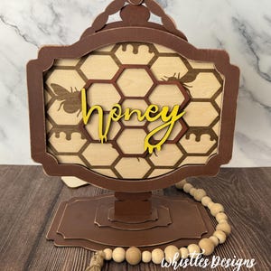 May include: A wooden honey-themed sign with a brown frame and base. The sign features a honeycomb pattern, bee silhouettes, and the word "honey" in yellow. A string of wooden beads and a tassel are at the base.