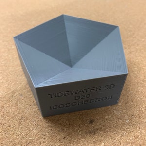 Puede incluir: Una torre de dados D20 de plástico gris con el texto "TIDEWATER 3D D20 ICOSOHERDRON" impreso en el lateral.