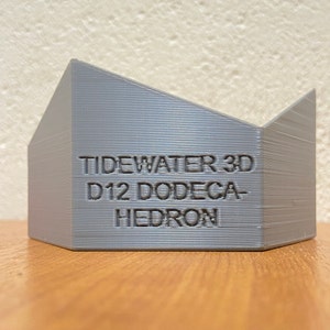 Puede incluir: Un dodecaedro D12 gris impreso en 3D con el texto "Tidewater 3D" en el lateral. La forma geométrica se muestra sobre una superficie de madera.