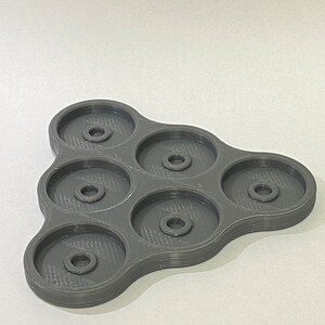 Pu&ograve; includere: Un vassoio di plastica grigia con sei scomparti circolari, ognuno con un piccolo foro al centro. Il vassoio ha la forma di un triangolo con bordi arrotondati.