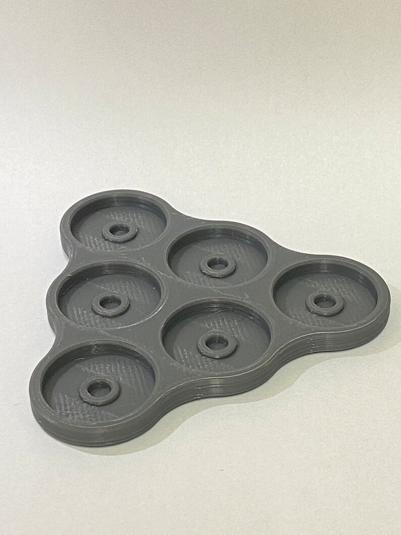 Pu&ograve; includere: Un vassoio di plastica grigia con sei scomparti circolari, ognuno con un piccolo foro al centro. Il vassoio ha la forma di un triangolo con bordi arrotondati.