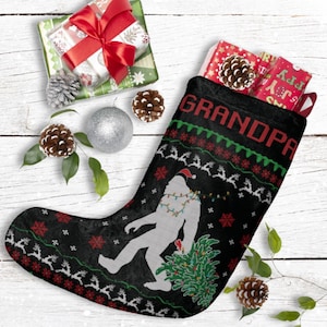 May include: Black Christmas stocking with the word "GRANDPA" in red. The stocking features a Bigfoot design wearing a Santa hat and Christmas lights, holding a Christmas tree. The stocking is filled with wrapped gifts and pine cones.
