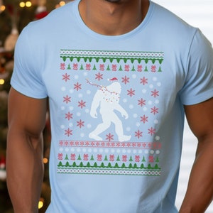 May include: Light blue t-shirt featuring a festive design. The shirt showcases a white Bigfoot silhouette wearing a Santa hat and string lights, surrounded by snowflakes and Christmas tree patterns. The design is in red, green, and white.
