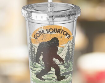 May include: A clear plastic tumbler with a lid and straw featuring a graphic of a dark brown Bigfoot silhouette walking through a forest landscape with a yellow sun in the background and the text GONE SQUATCH'N printed in a curved font above.