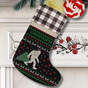 May include: A black Christmas stocking with a Bigfoot design carrying a Christmas tree. The stocking has a checkered top and the word "GRANDPA" in green. A red and white swirled lollipop and a yellow gift are inside the stocking. The stocking is hanging on a fireplace mantel.