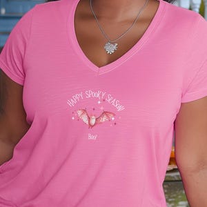 May include: A woman wearing a pink v-neck t-shirt with a bat graphic and the text "Happy Spooky Season". A Rottweiler puppy, pumpkins, and sunflowers are also visible. A tumbler with a bat graphic is on the right.